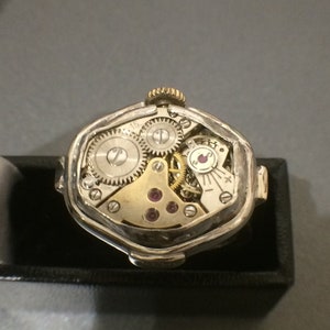 May include: A silver and gold watch with a unique octagonal shape. The watch is open, revealing the intricate inner workings of the mechanism.