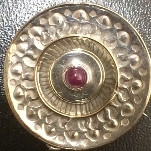 May include: A silver circular pendant with a raised, concentric circle design. The center circle features a small, round, pink gemstone.