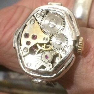 May include: A silver watch with a unique, octagonal-shaped case. The watch face is open, revealing the intricate inner workings of the movement. The watch has a gold-colored crown and several small, ruby-colored jewels.