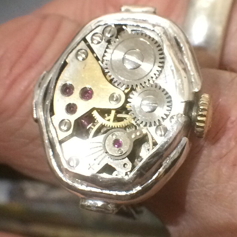 May include: A silver watch with a unique, octagonal-shaped case. The watch face is open, revealing the intricate inner workings of the movement. The watch has a gold-colored crown and several small, ruby-colored jewels.