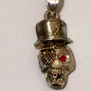 May include: A silver-toned metal pendant shaped like a skull wearing a top hat. The skull features a textured gold-toned eye patch over one eye, a small red gemstone in the other eye socket, and a tiny red gemstone accent on the teeth area.