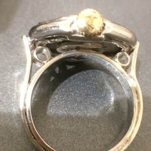 May include: A silver ring with a round, clear glass top and a gold-colored crown on top. The ring is designed to be a locket.