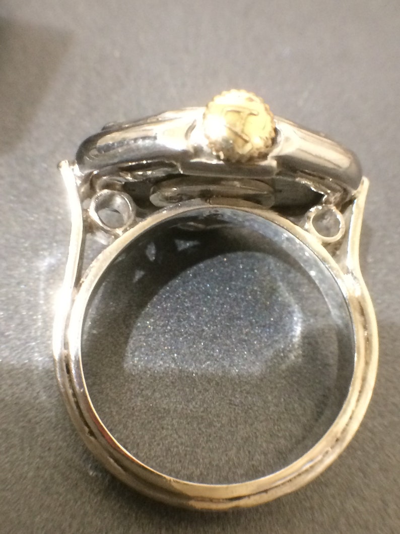 May include: A silver ring with a round, clear glass top and a gold-colored crown on top. The ring is designed to be a locket.