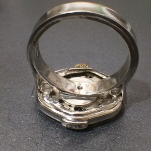 May include: A silver ring with a watch face embedded in the top. The watch face is visible through a clear glass cover. The ring is designed to be worn on the finger. The watch face has a silver band around the edge and a silver hand in the center. The ring is made of metal and has a silver finish.