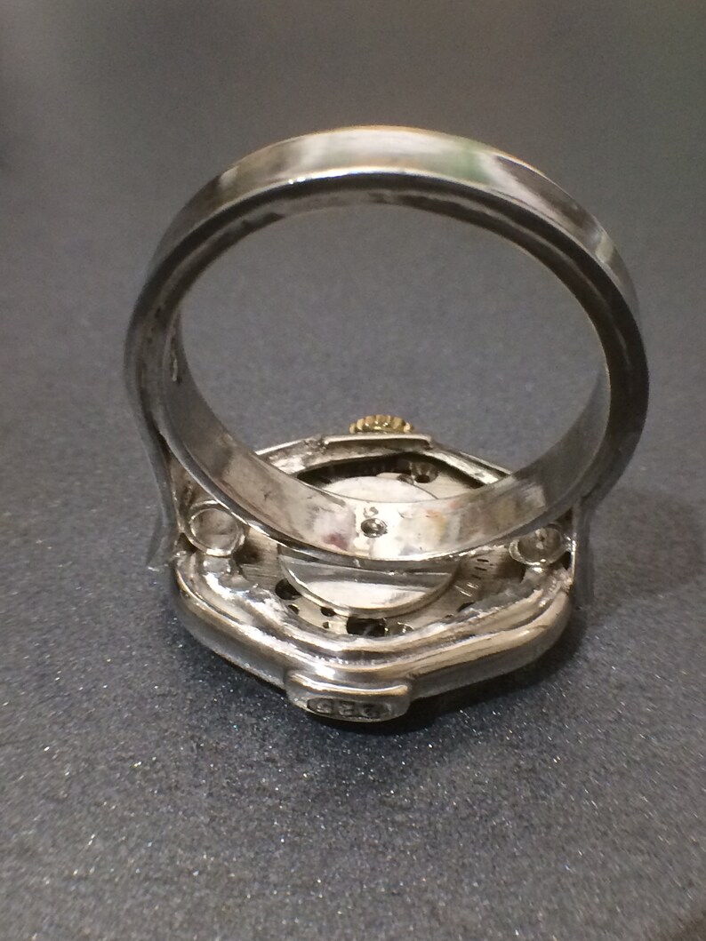 May include: A silver ring with a watch face embedded in the top. The watch face is visible through a clear glass cover. The ring is designed to be worn on the finger. The watch face has a silver band around the edge and a silver hand in the center. The ring is made of metal and has a silver finish.