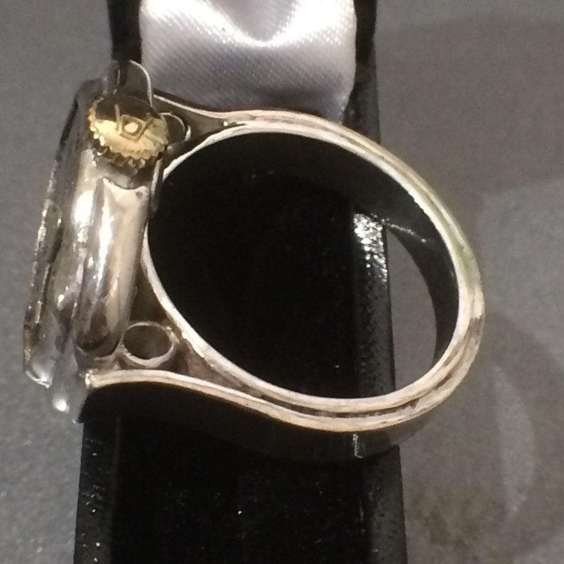May include: A silver ring with a round watch face attached to the top. The watch face has a gold rim and a small triangle at the top.