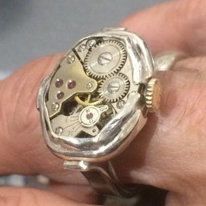 May include: A silver ring with a watch face embedded in the top. The watch face is exposed, showing the inner workings of the watch, including gears and a small ruby-colored jewel.