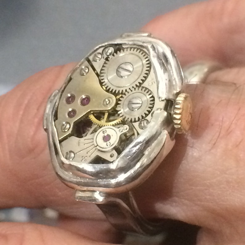 May include: A silver ring with a watch face embedded in the top. The watch face is exposed, showing the inner workings of the watch, including gears and a small ruby-colored jewel.