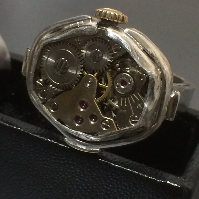 May include: A silver ring with a watch face embedded in the top. The watch face is open and shows the inner workings of the watch, including gears and a small ruby.