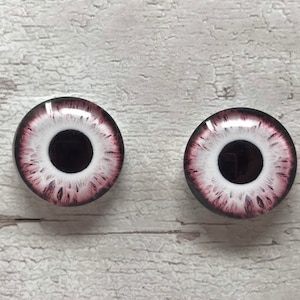 May include: Two white glass eyes with black pupils and pink detailing. The eyes are round and have a glossy finish.