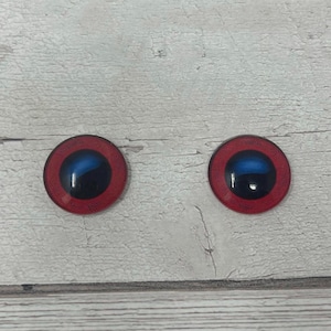 May include: Two round glass doll eyes. Each eye features a red outer ring, a dark blue centre, and a black pupil. The eyes are set against a light wood grain background.