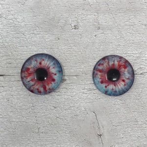 May include: Two realistic-looking glass eyes with red and white splatter patterns. The eyes have black pupils and are set against a white wooden background.