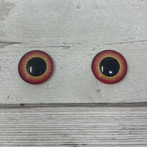 May include: Two round glass doll eyes featuring a black pupil and a red and yellow iris. The eyes are set against a light wood grain background.