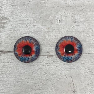 May include: Two round, colourful glass eyes with black pupils and red, blue and white irises. The eyes are on a white wooden background.
