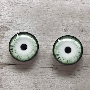 Op de afbeelding: Twee ronde, groene en witte glazen ogen met zwarte pupillen. De ogen zijn voor knutselen en poppen maken.