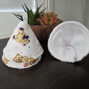 May include: White fabric nursing pads and a matching nursing cover with a teddy bear pattern. The cover is cone-shaped with a loop for hanging. The pads are round and made of a soft, absorbent material.