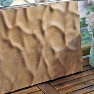 Puede incluir: Una pieza de arte rectangular de madera con una superficie tallada y ondulada. La madera tiene un color marr&oacute;n claro y un patr&oacute;n de vetas natural. La pieza se exhibe al aire libre, apoyada contra una barandilla.