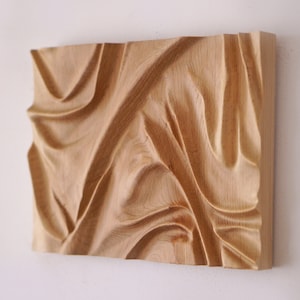 Op de afbeelding: Een houten wandsculptuur met een vloeibaar, abstract ontwerp. De sculptuur is gesneden uit lichtbruin hout en heeft een gladde, gepolijste afwerking.