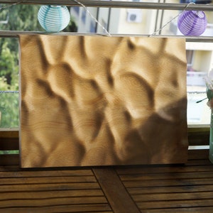 Puede incluir: Un panel rectangular de madera clara con una superficie texturizada, que se asemeja a dunas de arena, se exhibe al aire libre. El panel est&aacute; rodeado de objetos decorativos, incluyendo faroles de papel y un arreglo floral.