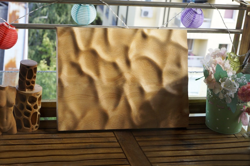 Puede incluir: Un panel rectangular de madera clara con una superficie texturizada, que se asemeja a dunas de arena, se exhibe al aire libre. El panel est&aacute; rodeado de objetos decorativos, incluyendo faroles de papel y un arreglo floral.