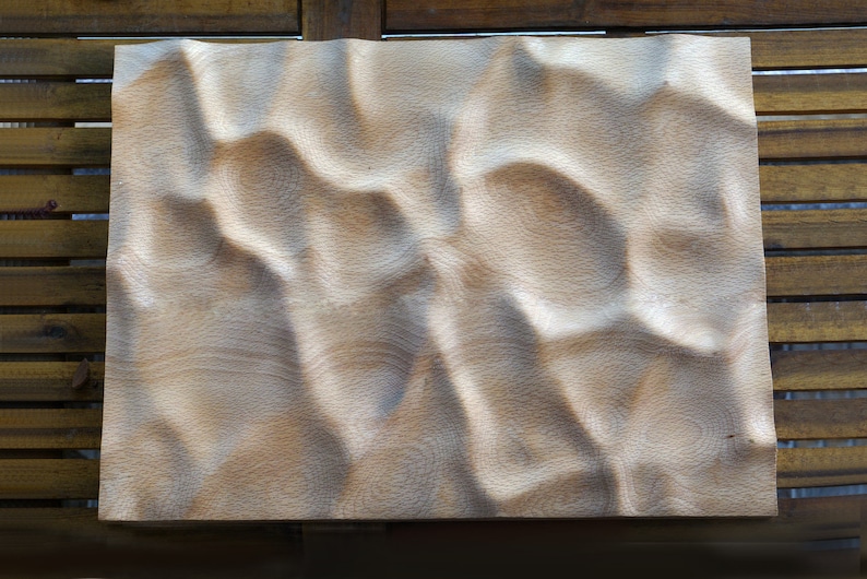 Puede incluir: Una obra de arte rectangular de madera con una superficie tallada y ondulada. La veta de la madera de color claro es visible, y la pieza est&aacute; colocada sobre un fondo de listones de madera. El tallado crea un patr&oacute;n similar a las olas.