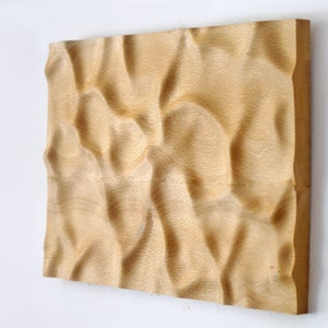 Puede incluir: Una obra de arte mural rectangular de madera con una superficie tallada y ondulada. La veta natural de la madera es visible, y la pieza est&aacute; montada en una pared blanca. La obra de arte es de color marr&oacute;n claro.