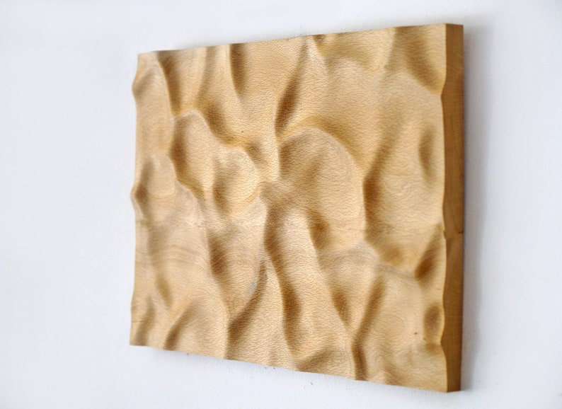 Puede incluir: Una obra de arte mural rectangular de madera con una superficie tallada y ondulada. La veta natural de la madera es visible, y la pieza est&aacute; montada en una pared blanca. La obra de arte es de color marr&oacute;n claro.