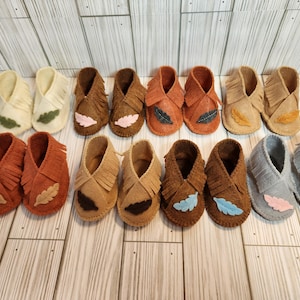May include: A collection of baby moccasin-style shoes in various colors, including cream, brown, and gray. Each pair features decorative leaf or feather accents and fringe detailing. The shoes are arranged on a wooden surface.