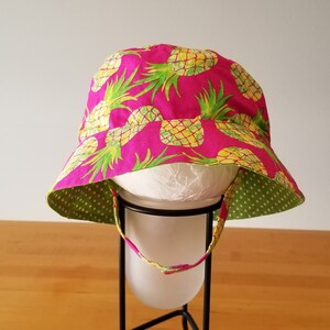 May include: A pink sun hat with a pineapple print and green polka dot lining. The hat has a wide brim and a chin strap.