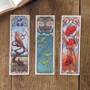 Op de afbeelding: Drie rechthoekige bladwijzers met Art Nouveau-ontwerpen. Eén toont een vogel op een tak, een ander een libelle en waterlelies, en de derde, rode klaprozen. Elk heeft een decoratieve rand en een illustratie met een natuurthema.