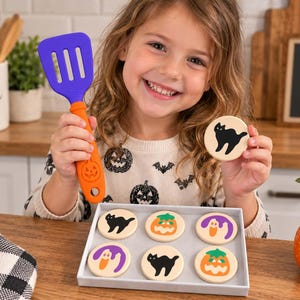 Peut inclure: Une spatule en silicone violette et orange avec un manche gaufré en forme de citrouille et un ensemble de six disques en bois ronds avec des motifs d'Halloween imprimés, dont des chats noirs, des citrouilles orange et des fantômes violets.