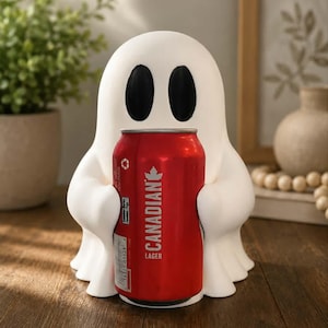 May include: A white ghost-shaped figurine with two black oval eyes, holding a red aluminium can of Canadian Lager. The ghost has a smooth, rounded form with small arms wrapped around the beverage container, which features white text and logos.