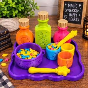 May include: A purple tray holding a purple cauldron, three colourful bottles with tan caps, two small cups, a yellow star-topped wand, and a yellow spoon. The cauldron and bottles feature embossed symbols like moons, stars, and leaves. The cauldron and spoon contain small yellow, blue, and pink plastic shapes.