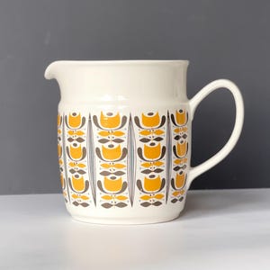 Könnte beinhalten: Cremefarbener Keramikkrug mit einem sich wiederholenden Muster aus gelben und braunen Blumenformen. Der Krug hat einen gebogenen Griff und einen Ausguss zum Ausgießen. Ein Küchenartikel im Vintage-Stil.
