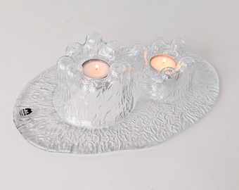 May include: A clear glass candle holder with two candle cups. The candle holder has a textured surface and is shaped like a wave. The candle cups are filled with lit tea lights.