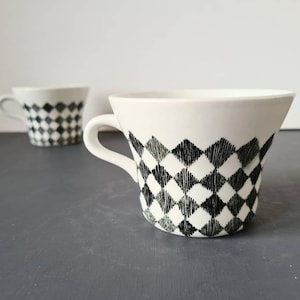 Op de afbeelding: Twee witte keramische koffiekopjes met een zwart-wit ruitpatroon. De kopjes hebben een uitlopende vorm en een handvat.