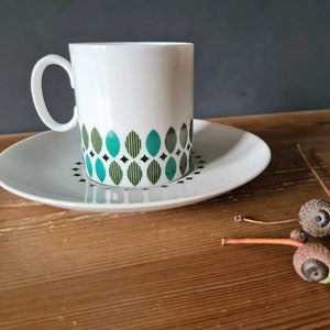 Könnte beinhalten: Eine weiße Kaffeetasse und Untertasse aus Keramik mit einem geometrischen Muster in Grün und Schwarz. Die Tasse hat einen abgerundeten Henkel. Die Untertasse ist schlicht weiß. Auf einer Holzoberfläche liegen vier Eicheln.
