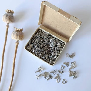 May include: A vintage box filled with silver-colored hook and eye closures, with some scattered outside the box. Two dried poppy seed heads stand to the left. The box is beige and open, revealing the contents.