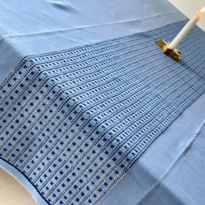 Puede incluir: Un mantel azul claro con un borde bordado en azul oscuro detallado. Una vela blanca encendida se encuentra en un soporte dorado sobre la mesa. El mantel tiene un diseño estampado.