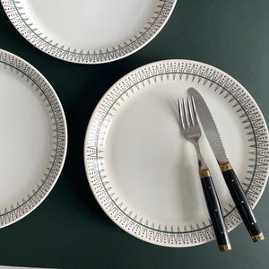 May include: White ceramic plates with a green and black geometric border design. A fork and knife with black handles and gold accents rest on one plate. The plates are on a dark green surface.