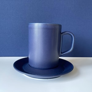 Puede incluir: Una taza de café de cerámica azul oscuro con un platillo a juego. La taza tiene una forma simple y cilíndrica con un asa redondeada.