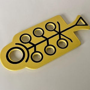 Può includere: Un sottopentola in ceramica gialla con un design astratto nero. Il sottopentola ha una forma arrotondata con una grande apertura circolare e sei aperture circolari più piccole.