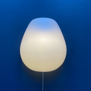 Op de afbeelding: Een witte en blauwe, matglazen lamp met een afgeronde, traanvormige vorm. De lamp is aan een muur gemonteerd en heeft een wit snoer.