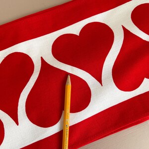 May include: Red fabric with a white border of repeating heart shapes. A yellow pencil is lying on top of the fabric.