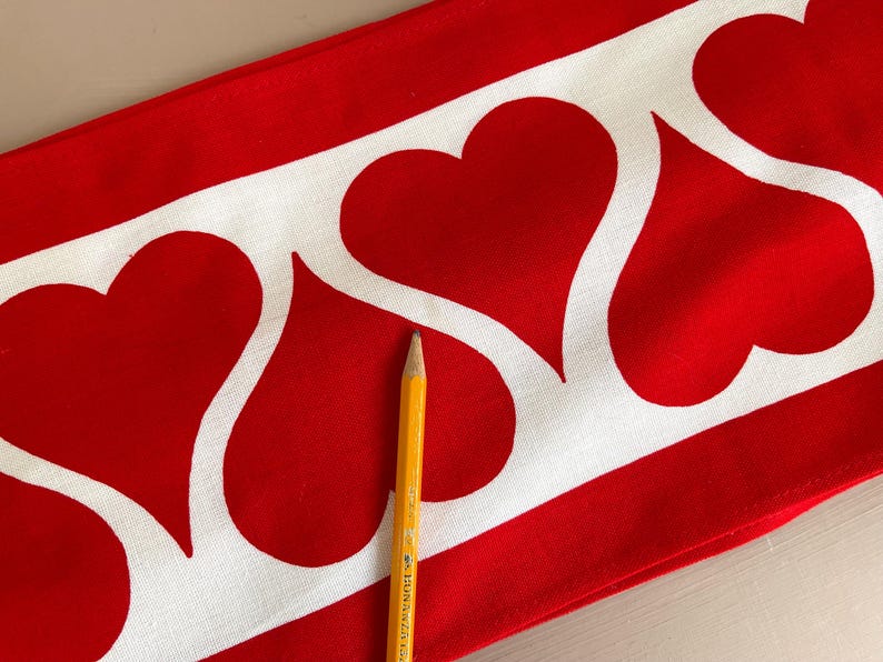 May include: Red fabric with a white border of repeating heart shapes. A yellow pencil is lying on top of the fabric.