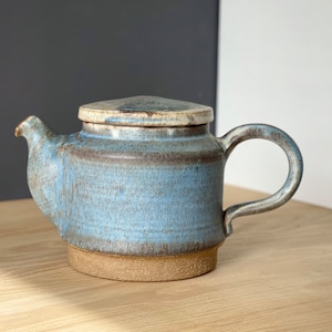 Può includere: Una piccola teiera in ceramica blu con un coperchio e un manico marrone. La teiera ha una glassa blu chiaro con accenti marroni.