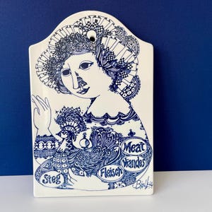 Puede incluir: Placa de pared rectangular de cerámica blanquecina con una ilustración azul de una mujer y un pájaro. La mujer lleva un sombrero y un vestido decorativos. La placa tiene las palabras "Steg", "Fleisch" y "Meat" en azul.