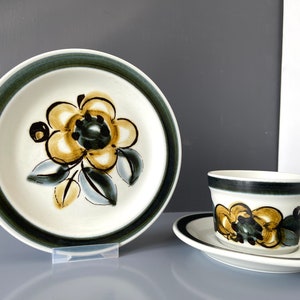 以下が含まれることがあります： 花柄のヴィンテージティーカップとソーサーのセット。カップとソーサーは白地に緑と茶色の縁取りが施され、黄色い花に茶色と青色のアクセントが添えられています。