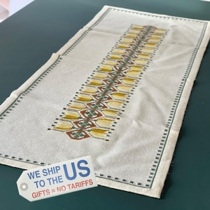 Op de afbeelding: Crèmekleurige rechthoekige tafelloper met een centraal geborduurd ontwerp in gele, bruine en groene geometrische patronen. De loper heeft een groene rand met een stippenpatroon. De tekst "WE SHIP TO THE US GIFTS = NO TARIFFS" staat op een label.