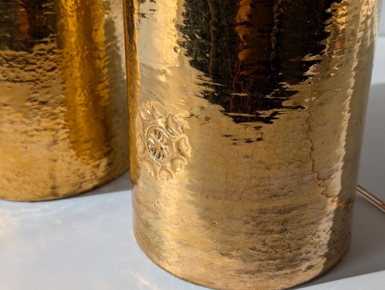 May include: Two gold metal table lamps with a hammered finish and a decorative floral design. The lamps have a distressed look.